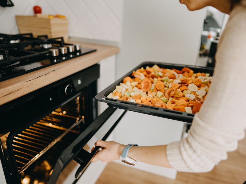 A imagem mostra uma mulher, que veste uma blusa com creme, abaixando-se em direção ao forno, abrindo o mesmo com a mão esquerda e, com a direita, segurando uma bandeja preta com legumes como cenoura, batata e afins. No interior no forno, podemos ver a luz amarela acesa. Acima dele, temos uma bancada com madeira clara e fogão cooktop, com botões prateados.