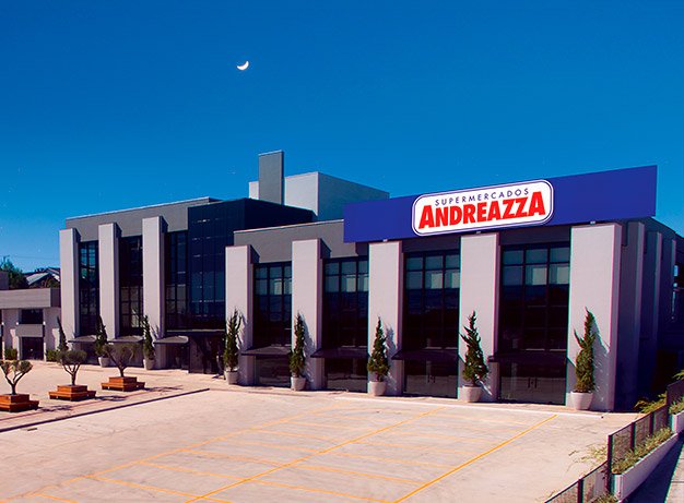 a foto mostra a frente de uma loja dos mercados Andreazza. O espaço do estacionamento, que é amplo, está vazio e possui diversas árvores dispostas no ambiente. A estrutura intercala colunas e grandes janelas de vidro e, acima dela, temos o painel com o logo do mercado, em azul, branco e vermelho.