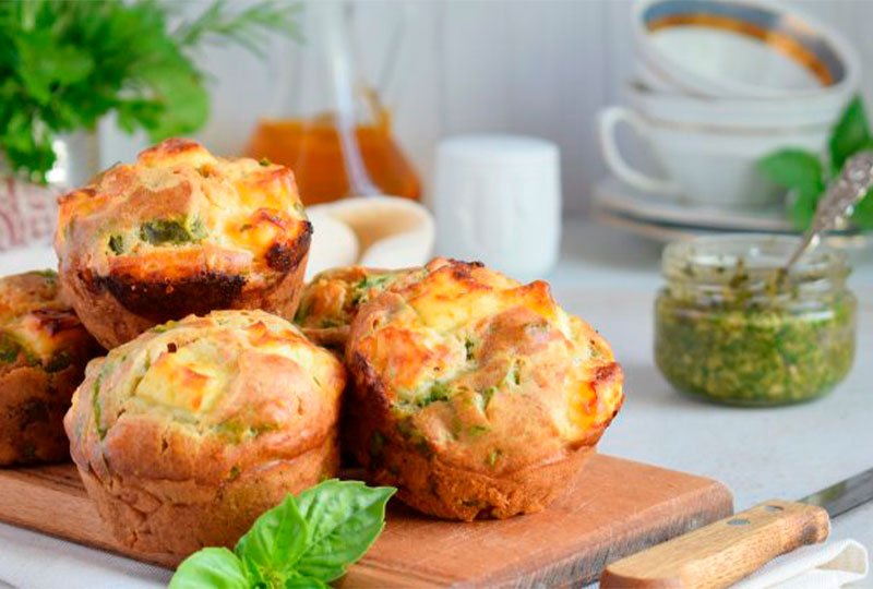 Foto de vários cupcakes salgados em cima de uma tábua de cortar, com uma faca ao lado com cabo de madeira. Ao fundo, também temos um recipiente com molho verde e uma colher dentro, tigelas e canecas e uma jarra com líquido alaranjado dentro.