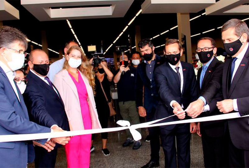 a imagem mostra pessoas puxando uma fita cerimonial que marca a inauguração de uma nova loja. Do lado direito, temos três homens, de terno e máscara, puxando um lado da fita. No lado direito, temos dois homes, também de ternos escuros e máscaras, e uma mulher, com roupas rosas, que puxam o outro lado da fita. Ao fundo, o público acompanha o ato, com câmeras registrando o momento.