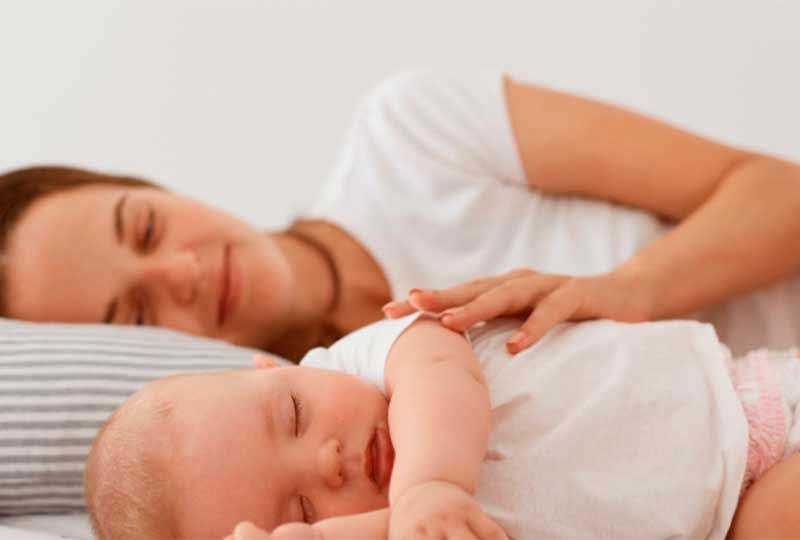 a imagem mostra uma mãe, de cabelos escuros e camiseta branca, deitada na cama com seu filho recém-nascido à frente. Ambos dormem tranquilamente, de lado, com a mãe sorrindo e com a mão esquerda posicionada suavemente sobre o corpo do filho.