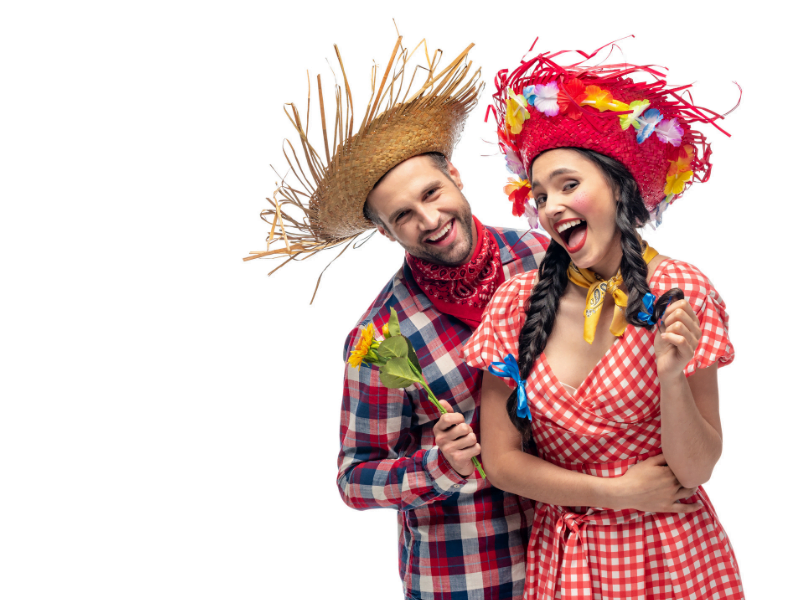 a imagem mostra um casal caracterizado com as roupas típicas das festas juninas. O rapaz usa camisa xadrez, em azul, vermelho e branco, um chapéu de palha na cabeça, lenço vermelho no pescoço, segura um ramalhete de flores, e sorri olhando para a câmera. A moça, que está próxima ao rapaz, tem cabelos escuros e veste um vestido xadrez em vermelho e branco, com um lenço amarelo amarrado no pescoço. Tem duas trancinhas no cabelo, e na ponta delas, possui lacinhos azuis. Ela também usa um chapéu vermelho com enfeites diversos e sorri para a câmera.