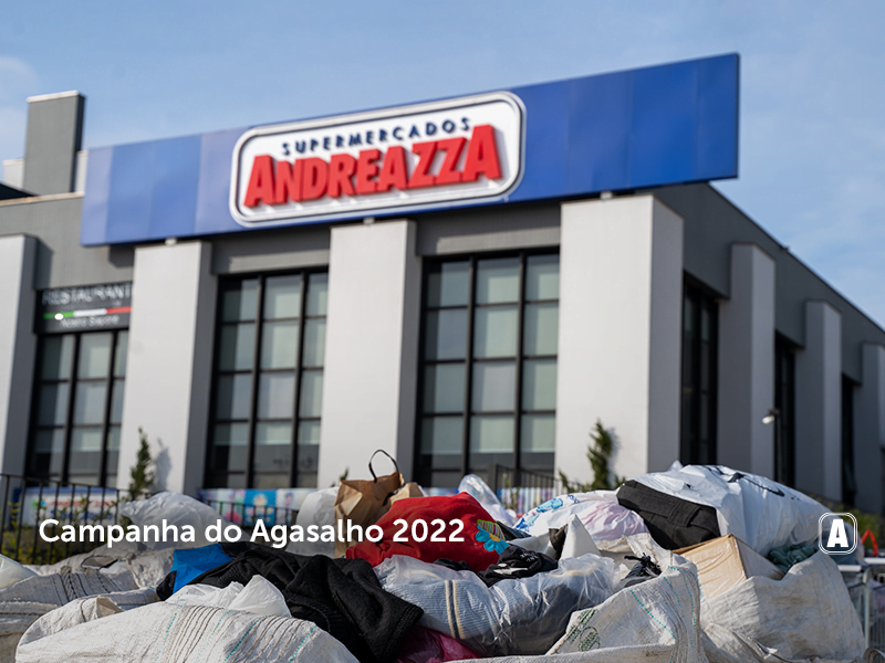 na foto, vemos a fachada do Supermercado Andreazza durante o dia, com as janelas abaixo do letreiro. Mais próximo da câmera, em primeiro plano, temos diversas sacolas de roupas, com destaque para peças vermelhas, pretas e azuis.