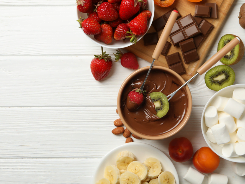 a imagem mostra uma cumbuca de chocolate com dois garfos de fondue, com um morango e um kiwi na ponta de cada um. Ao redor, temos um prato com bananas e uma cumbuca com marshmallows. Espalhado sobre a mesa há um kiwi cortado ao meio, morangos, amêndoas, ameixas e marshmallows. Há também uma tábua com pedaços de chocolate em barra.