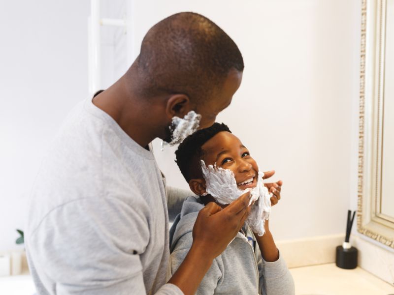 A imagem mostra pai e filho em frente ao espelho de um banheiro. Ambos são afrodescendentes e estão no processo de fazer a barba. A criança, mais baixa que o pai, olha para cima sorrindo, enquanto o pai passa creme de barbear no rosto do filho. Ambos vestem casacos cinzas, mas o da criança possui capuz. O banheiro em que estão é branco, com paredes e pias em detalhe de mármore. A moldura do espelho, em frente aos dois, é de madeira e num tom um pouco mais escuro que o da parede.