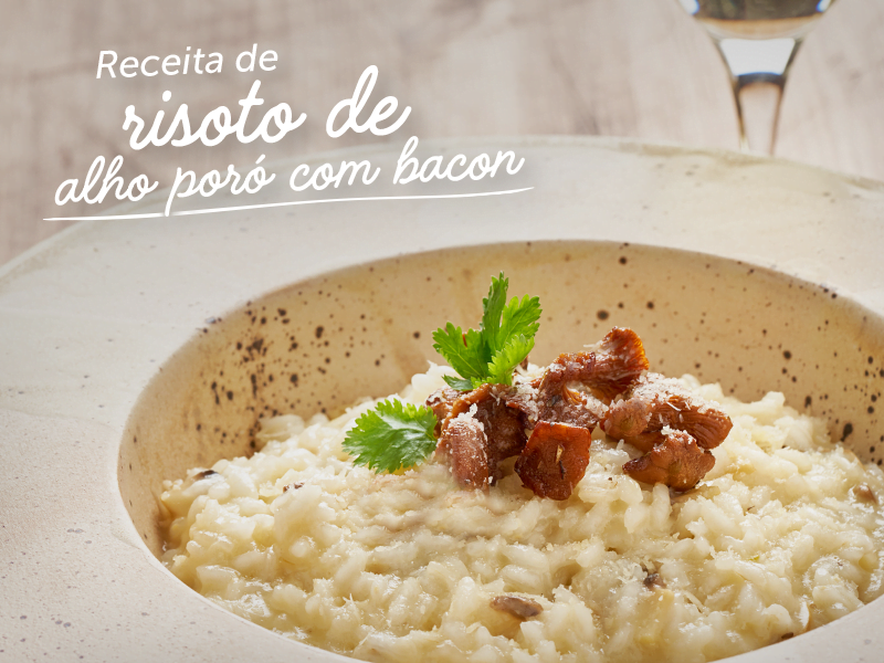 a imagem mostra um prato branco colocado em cima de uma mesa de madeira clara. Dentro do prato, vemos um risoto de grãos brancos com bacon e folhas verdes no topo. Ao redor do prato, na parte inferior, temos uma pequena tigela com arroz grosso e folhas verdes em cima de um pano. Na parte superior do prato, uma taça com vinho branco e uma parte da garrafa exibida.