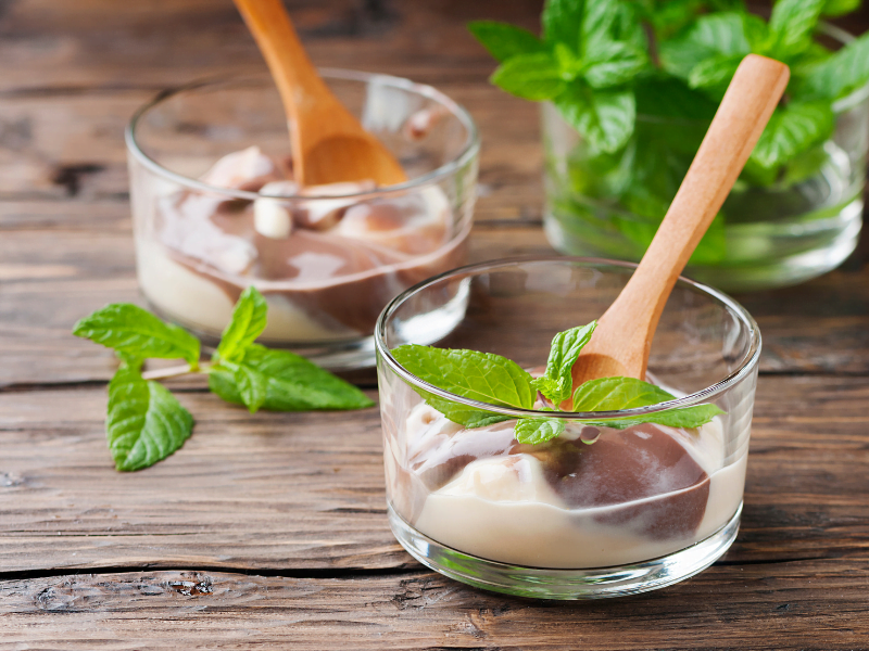 A imagem mostra três potes cilíndricos transparentes, pequenos, em cima de uma superfície de madeira escura. Nos dois potes mais próximos, há o mousse com chocolate, decorado com folhas de hortelã e pequenas colheres de pau. Ao fundo, no terceiro pote há água e folhas de hortelã dentro.