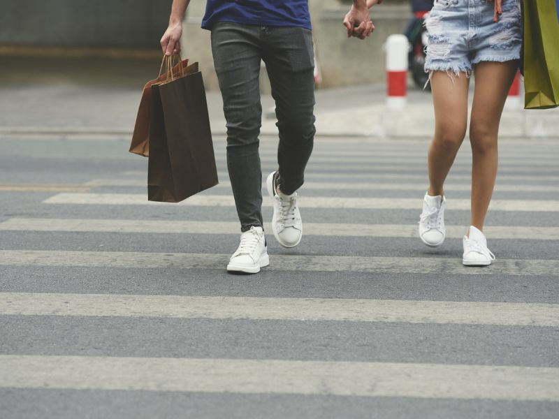 A imagem duas pessoas, de mãos dadas, atravessando a rua na faixa de segurança. A pessoa da esquerda veste sapatos brancos, calça escura e camiseta azul, e carrega duas bolsas de compras na mão direita. A pessoa à direita do observador também calça sapatos brancos, um shorts jeans claro e carrega uma bolsa no lado direito.