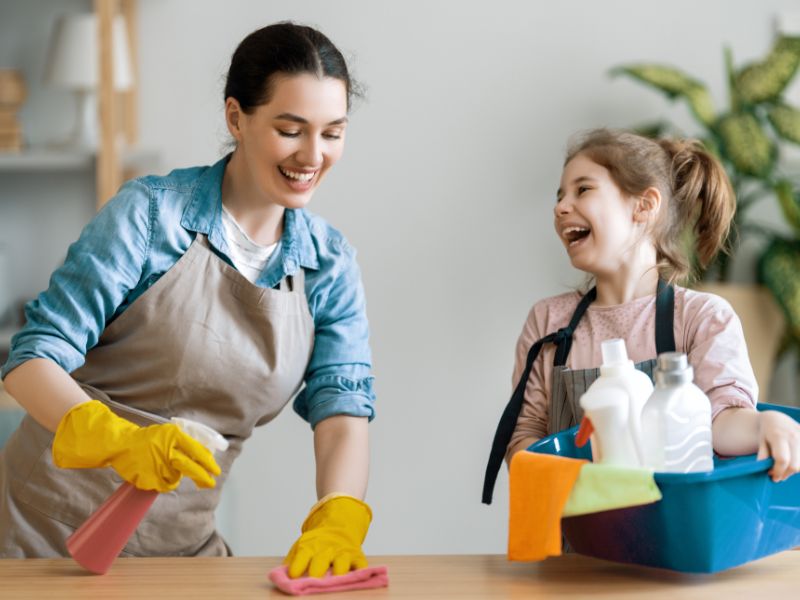 A imagem mostra mãe e filha. A mãe, tem cabelos escuros que estão presos em um coque. Ela usa camisa jeans azul-claro, e por cima dela, um avental marrom. Veste luvas de borracha amarelas, e enquanto ri, passa um pano em uma mesa de madeira de tom claro. A filha, na faixa dos 10 anos, tem cabelo loiro-escuro que está preso em um rabo de cavalo. Ela veste camiseta salmão e por cima, um avental. A filha está ao lado de sua mãe, olhando e rindo para ela, enquanto segura uma bacia com panos e produtos de limpeza.