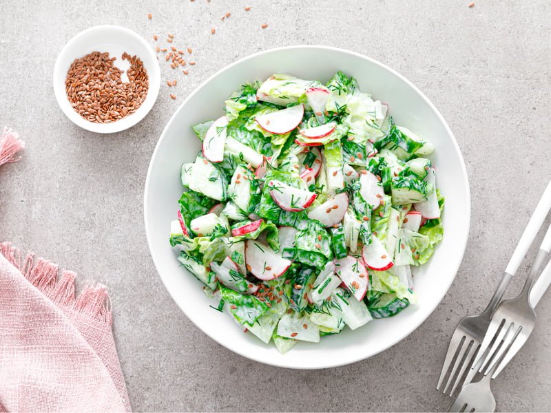 A imagem mostra um prato branco sobre uma superfície de granito clara. Dentro dele, vemos salada de folhas verdes com fatias de rabanetes, pepinos e salpicados com grãos pequenos. Todos os ingredientes estão cobertos com um molho branco. Ao lado direito do prato, vemos três garfos. No lado esquerdo, uma toalha rosa pastel e uma tigela pequena com grãos.