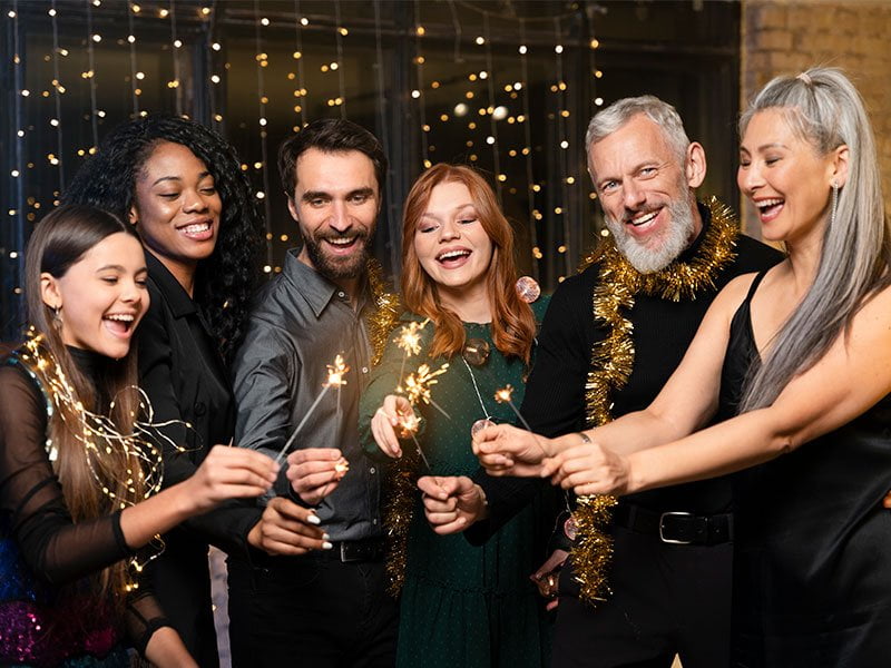 A imagem mostra um grupo de 6 pessoas reunidas, 4 mulheres e 2 homens, com todos segurando uma vela-faísca. A garota da esquerda possui cabelos escuros e pele clara, com luzes de Natal enroladas no pescoço; ao seu lado, uma mulher de cabelos escuros cacheados, pele morena e roupa preta; um homem de cabelos e barba pretos, com camisa cinza-escuro, próximo a uma mulher de cabelos ruivos e camisa verde; um homem de cabelos curtos e barba comprida brancos, com roupas pretas e um enfeite dourado no pescoço; uma mulher com feições asiáticas, de cabelos longos grisalhos e vestido preto.