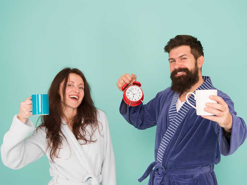 A imagem, com fundo azul-claro, mostra um casal com roupões, ambos de cabelos pretos. O homem veste roupão azul escuro, e a mulher um roupão branco. Ambos seguram xícaras nas mãos, como se estivessem recém acordados. O homem também segura um despertador vermelho na mão direita. O casal sorri enquanto olha para a câmera.