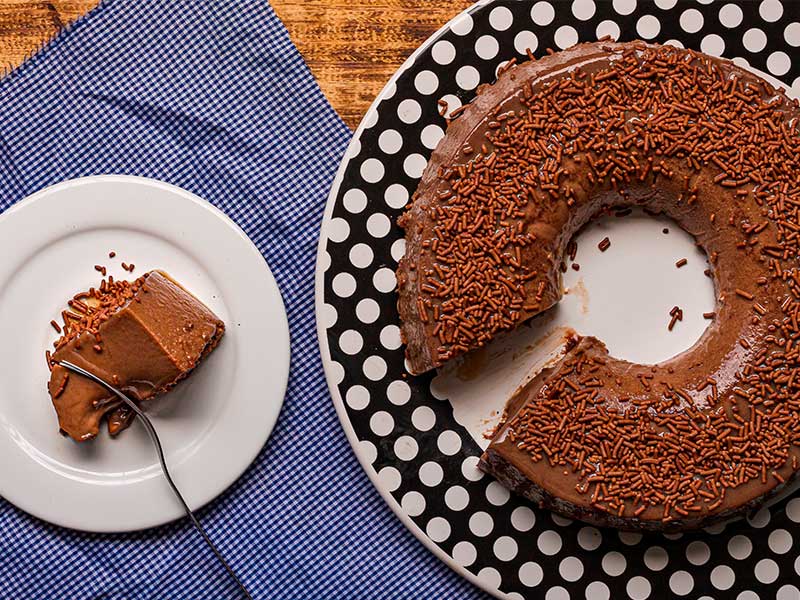 A imagem mostra uma foto, aérea, de um bolo de brigadeiro polvilhado. O bolo, que está em um prato com detalhes e bolinhas brancas, está cortado, com o pedaço do mesmo colocado sobre um prato branco, à esquerda do bolo.