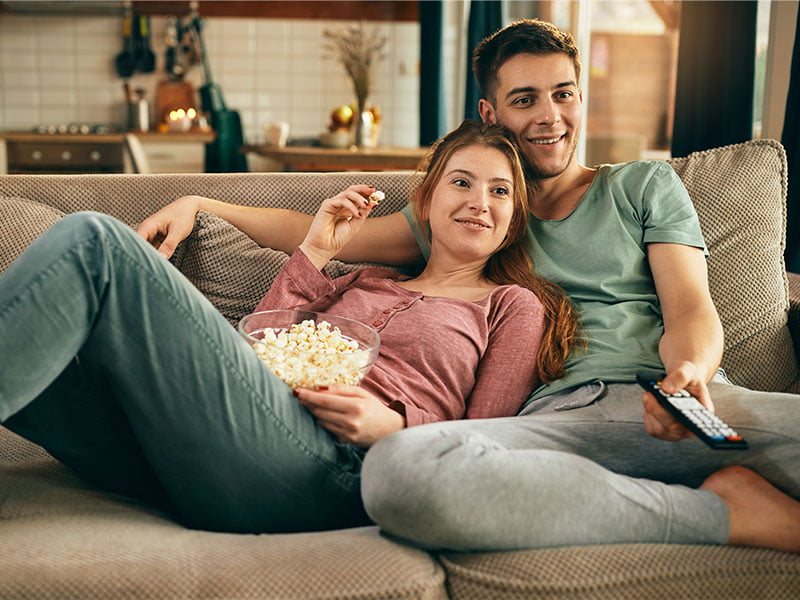 casal-sorridente-comendo-pipoca-e-assistindo-tv-enquanto-relaxa-no-sofa