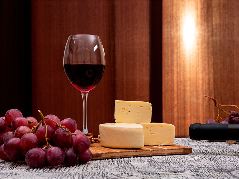 A imagem mostra uma mesa posta com cacho de uva, uma tábua com queijos e uma taça de vinho tinto, preenchida até a metade. No canto da foto, vemos o gargalo de uma garrafa ainda tampada, galhos de um segundo cacho de uva. O fundo mostra uma parede de madeira.