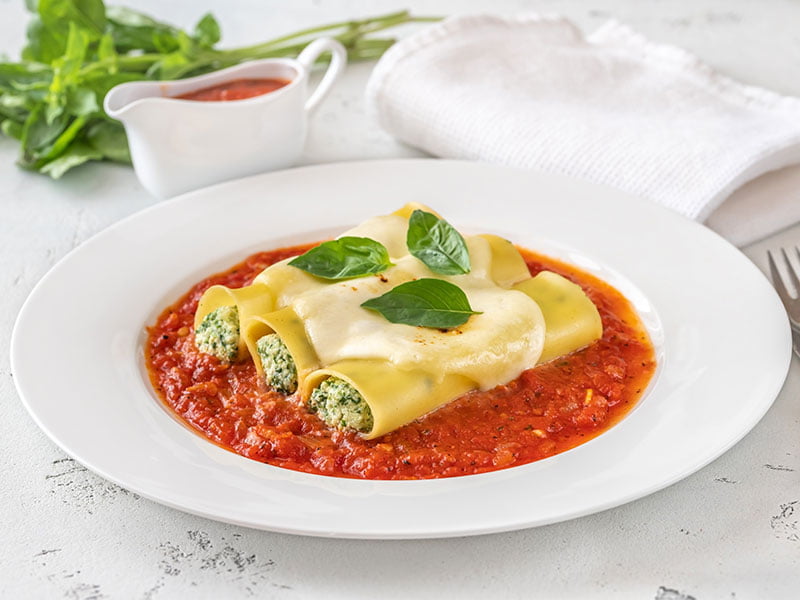 A imagem mostra três canelones com recheio de ricota colocados lado-a-lado em um prato branco com o fundo preenchido de molho vermelho. Os canelones estão decorados com um molho branco e 3 folhas no topo. Ao lado do prato, vemos os talheres: garfo e faca. Ao fundo da imagem, atrás do prato, vemos uma toalha branca dobrada, uma molheira com o mesmo molho vermelho do prato, e folhas verdes. Tudo isso está disposto acima de uma bancada branca com algumas marcas de desgaste/uso.