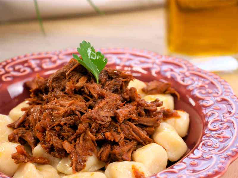 Na imagem, há um prato ornado com a cor bordô com uma porção de nhoques. O prato está sobre uma mesa e os nhoques estão temperados com ragu de carne. Na apresentação do prato, também é possível notar a presença de salsinha.