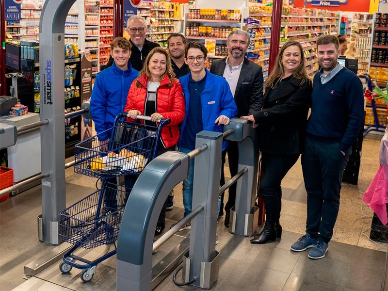 A imagem mostra 8 pessoas no corredor de um supermercado, passando por estrutura metálica. A mulher mais a frente do grupo, que usa um casaco vermelho, segura um carrinho de compras com dois compartimentos. Há alguns itens colocados no compartimento superior. Ao fundo, vemos as prateleiras do mercado.