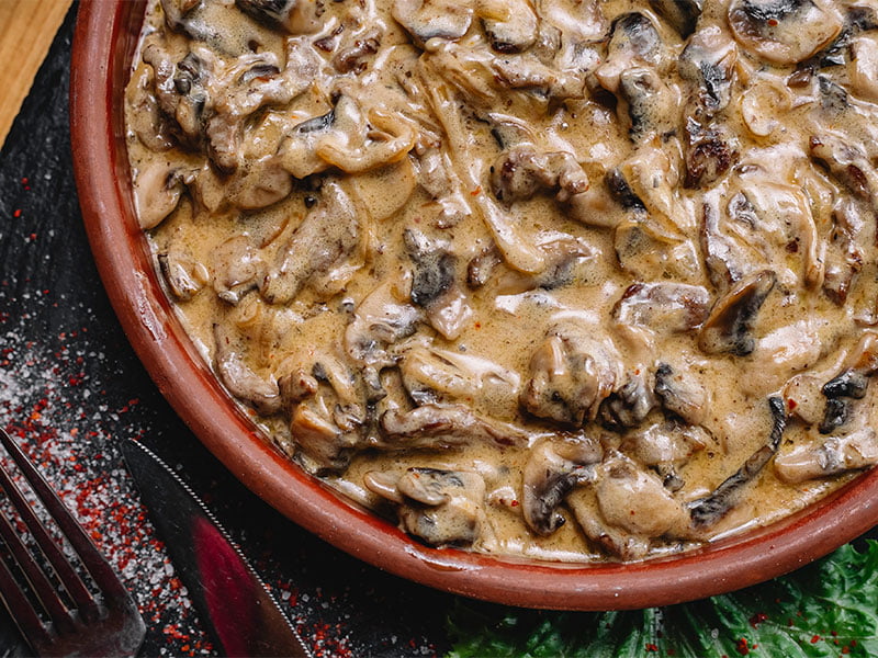 Sobre uma mesa de madeira e uma estrutura preta, vemos um prato vermelho que contém estrogonofe em seu interior. Na imagem, também há uma folha de salada ao lado do prato, decorando a apresentação, junto a um garfo e uma faca.