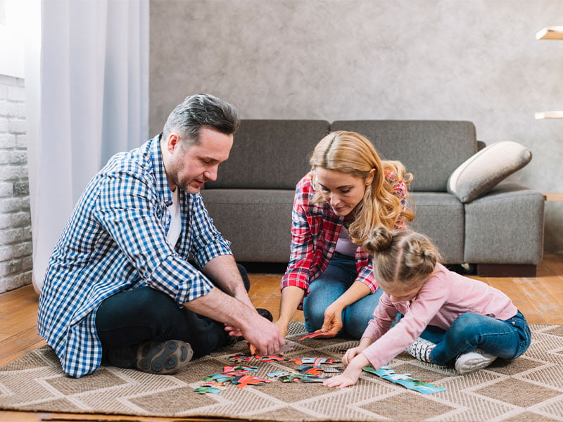 No chão da sala de uma casa, com um sofá ao findo, piso de madeira e parede cinza, uma família brinca. Na imagem, está o pai, a mãe e uma filha, brincando de montar quebra-cabeças.