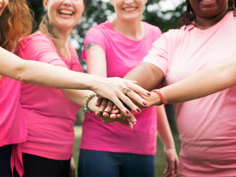 Na imagem, 4 mulheres de camiseta rosa estendem as mãos, com uma se sobrepondo a outra e demonstrando união. Apesar as imagem estar desfocada, é possível perceber que todas elas estão sorrindo!
