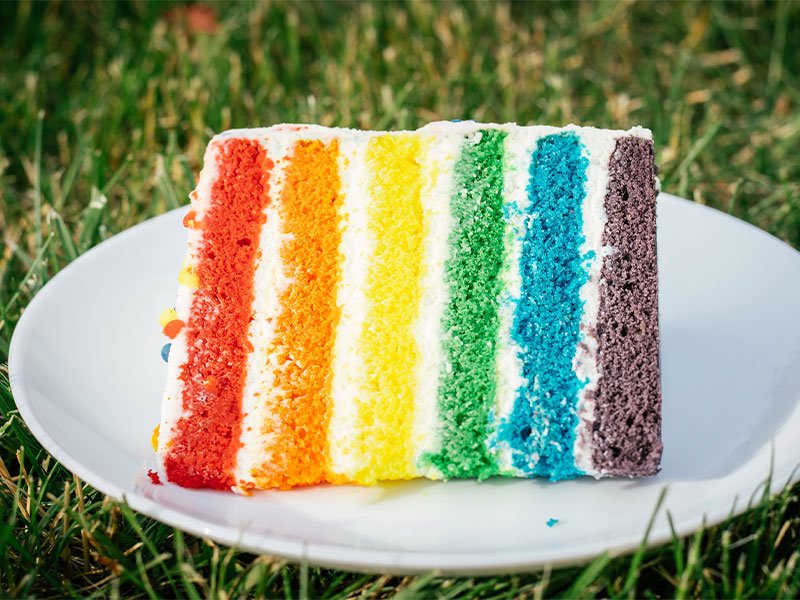 Na imagem, há uma fatia de bolo arco-íris, com as cores vermelho, laranja, amarelo, verde, azul e roxo. O pedaço de bolo está sobre um prato que está posicionado em um gramado.