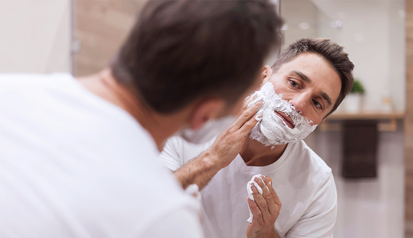 Na imagem, um homem de camiseta branca e tatuagem no braço se observa no espelho enquanto cobre a sua barba com creme para barbear.