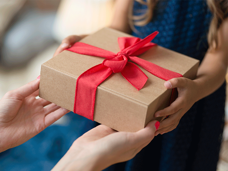 Na imagem, as mãos de uma menina recebem uma caixa de presente marrom, com um laço vermelho, das mãos de uma mulher. A imagem remete à troca de presentes em família durante o Natal.