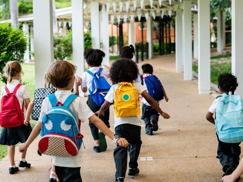 Um total de nove crianças está adentrando uma escola. Todos estão com mochilas nas costas.