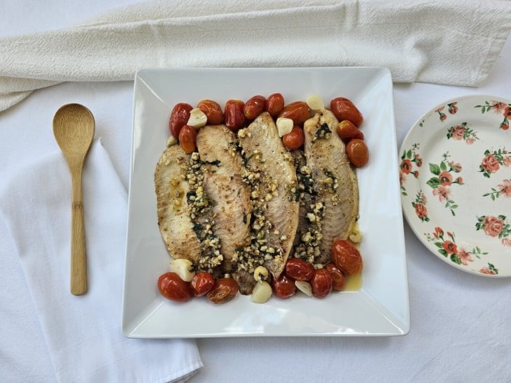 Sobre uma superfície branca, estão alguns utensílios de cozinha e um prato branco com filé de tilápia com tomate confit e molho de castanha.