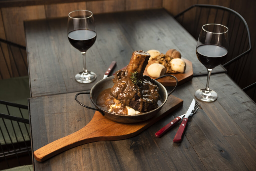 Mostra uma mesa com um prato de ossobuco e taças de vinho tinto.