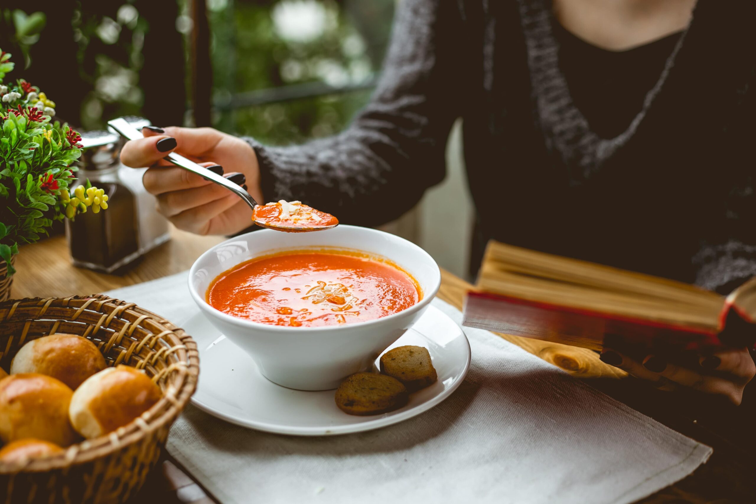 Mão feminina segurando colher diante de um prato rústico com sopa cremosa de tomate.