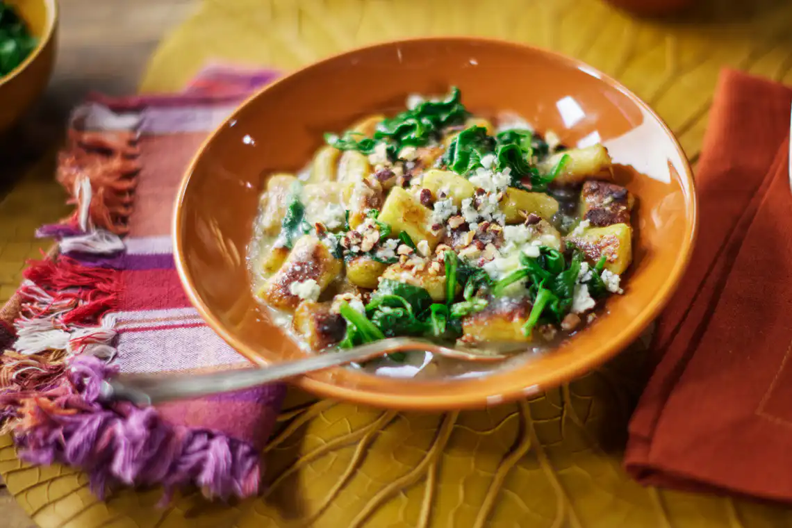 Prato de cerâmica contendo nhoques de batata dourados envolvidos em molho de manteiga, decorados com folhas de sálvia fresca e queijo ralado.