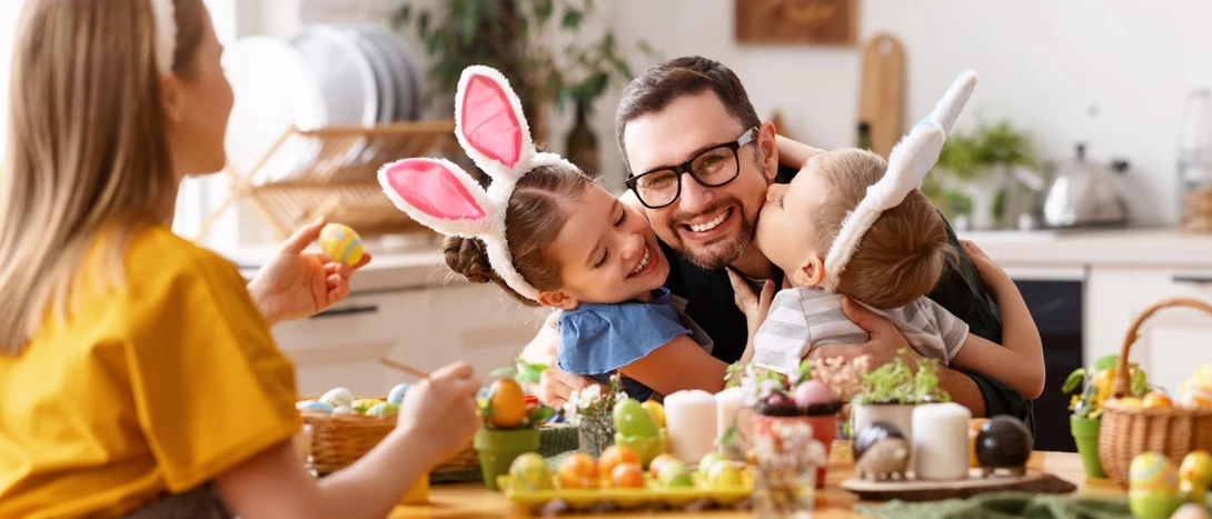 Duas meninas pequenas, usando tiaras com orelhas de coelho, abraçam o pai com entusiasmo, enquanto a mãe, sentada à mesa, pinta ovos de Páscoa com um pincel e tintas coloridas ao lado.